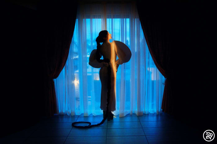 Woman in a flowing white dress standing in front of a window with white and blue curtains, illuminated by a bright light.