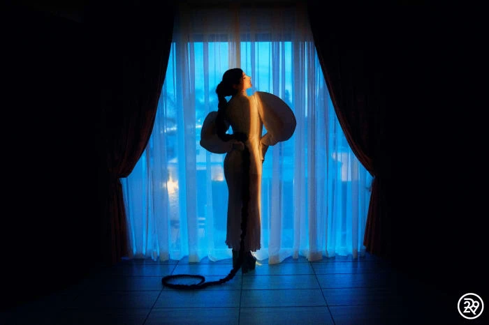 Woman in a flowing white dress standing in front of a window with white and blue curtains, illuminated by a bright light.