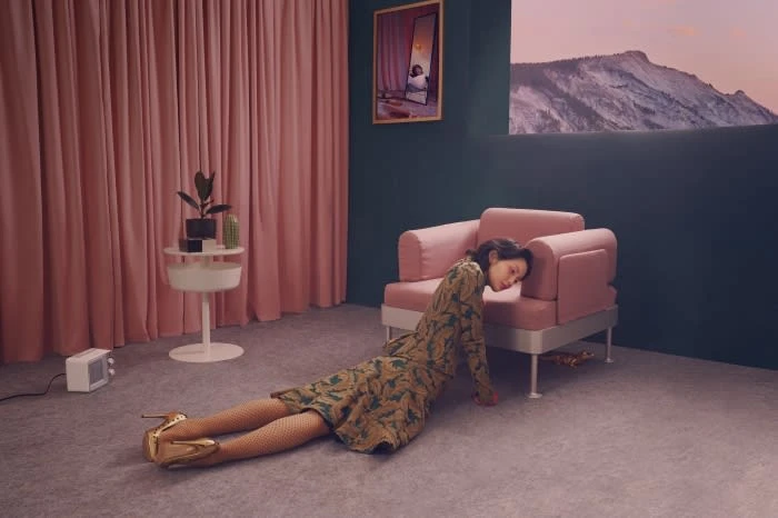 Woman in patterned dress and gold heels rests head on a pink couch in an indoor setting with pink curtains and mountain view.