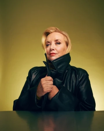Blonde woman wearing a dark leather coat with a high collar seated against a yellow-green background by photographer Michelle Watt