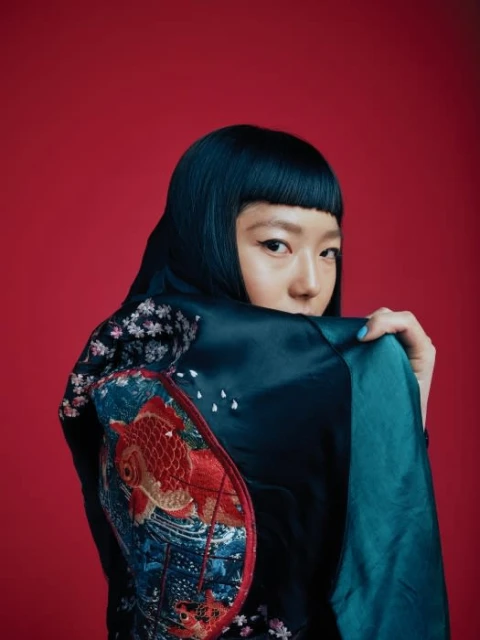 Portrait of a young Asian woman with black hair and a blue silk jacket with an embroidered goldfish and flowers on a red background.