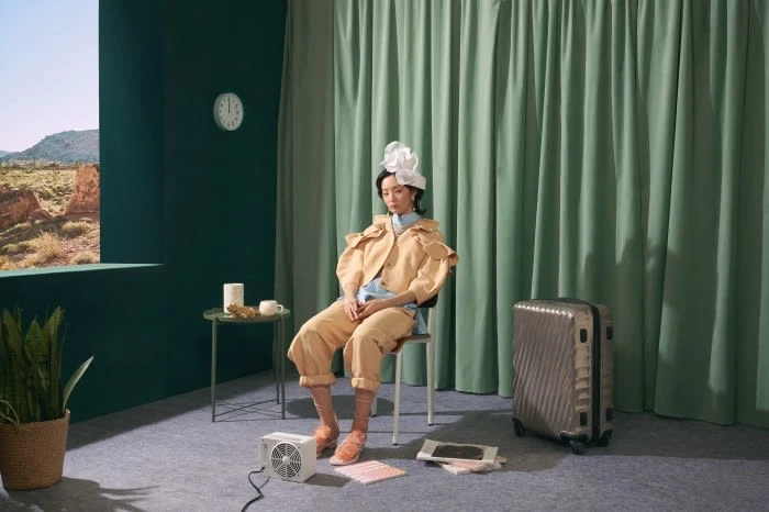 Woman in a tan suit and large white headpiece poses while seated on a chair in an indoor studio setting with a suitcase nearby.