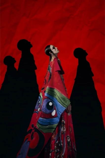 A fashionable woman in a red eye-print gown looks up against a matching red backdrop with silhouetted figures.