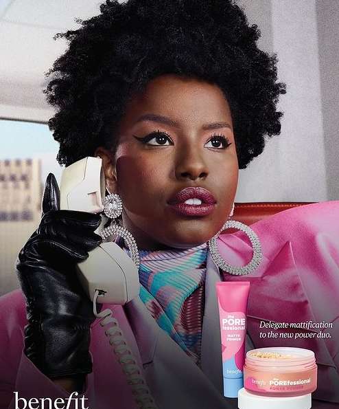 Beauty ad for Benefit Cosmetics features Black female model in pink blazer, holding retro telephone, and product display.