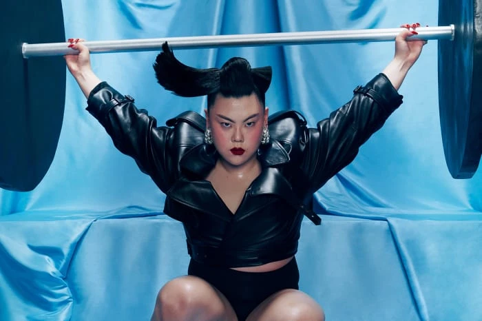 Adult lifting a barbell with theatrical makeup and a unique hairstyle in front of a light blue draped backdrop, studio shot.