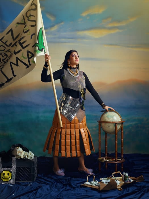 Woman holds climate protest banner with globe. She wears a skirt constructed from stacked leather and poses in an outdoor setting.
