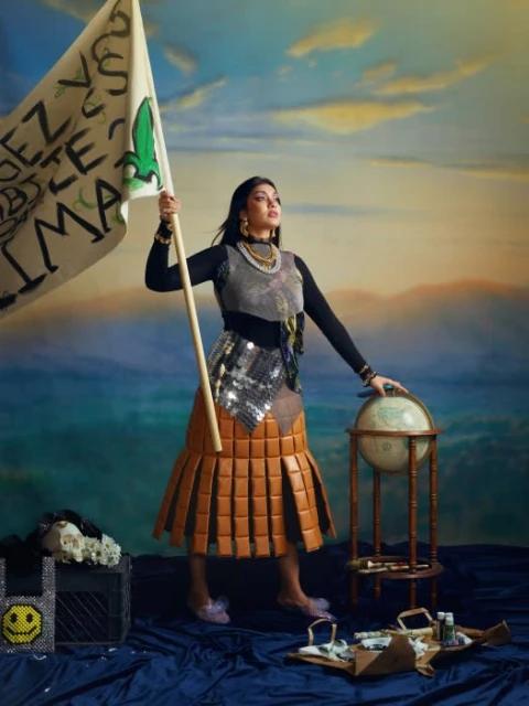 Woman holds climate protest banner with globe. She wears a skirt constructed from stacked leather and poses in an outdoor setting.