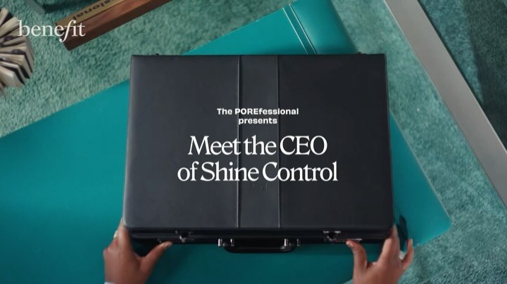 Eye-level shot of a black briefcase with white text, held by a person with manicured hands against a teal and carpeted backdrop.