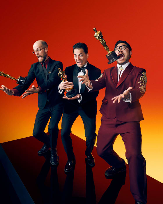 Three men in formal suits playfully pose with awards against a vibrant orange background, showcasing celebratory expressions and dynamic stances.