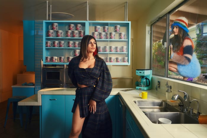 Woman in a dark plaid outfit poses in a light blue kitchen with another woman looking through the window in a colorful hat.