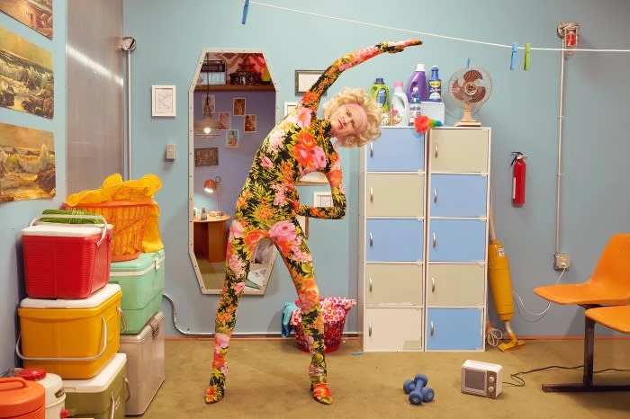 Woman in floral catsuit stretching in a brightly lit laundry room with colorful appliances, cleaning supplies, and vintage decor.