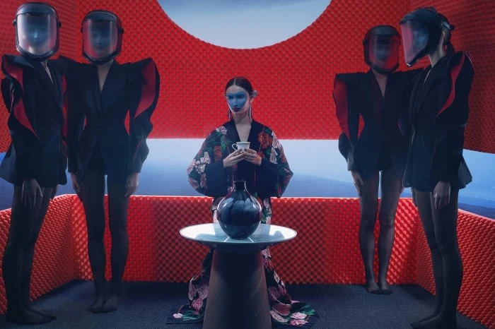 Artistic fashion portrait with a central model holding a teacup, flanked by helmeted figures in a room with textured red walls and blue light.