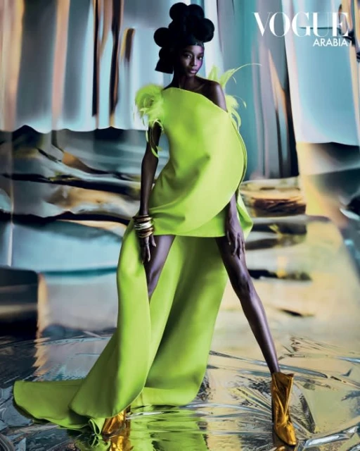Fashion editorial shot of a slender Black model wearing a lime-green gown with gold boots against a mottled silver and grey backdrop.
