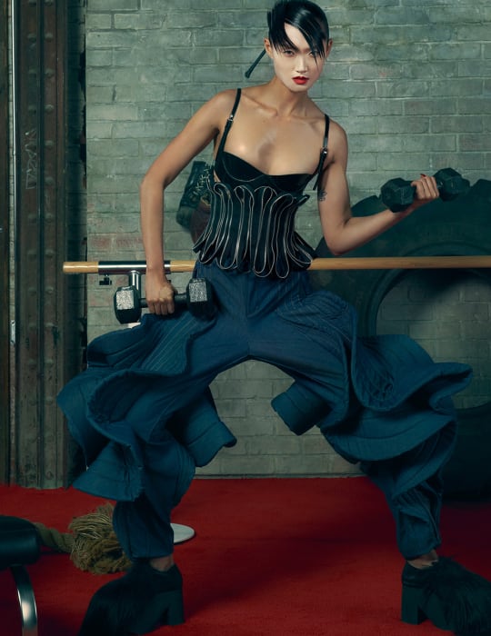 Athletic woman in editorial fitness fashion lifts dumbbells indoors, featuring textured brick background and dark blue ruffled pants.