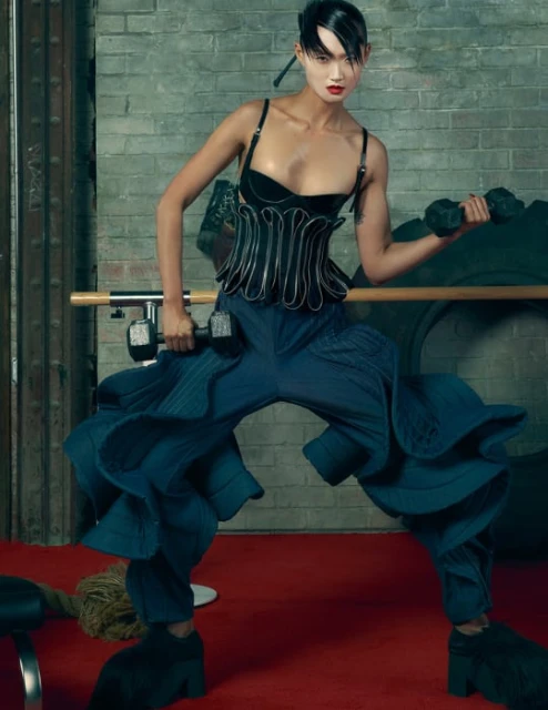Athletic woman in editorial fitness fashion lifts dumbbells indoors, featuring textured brick background and dark blue ruffled pants.