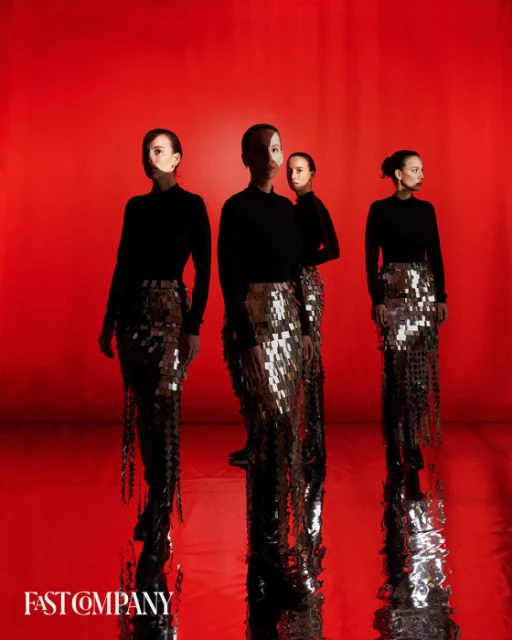 Four women in black tops and sequin skirts stand against a vivid red backdrop, reflecting off the shiny floor in a fashion-style studio shoot.