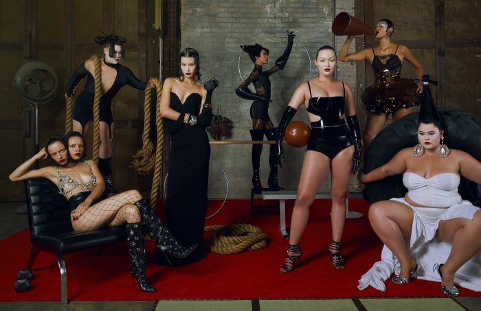 Group of seven fashion models in a studio setting wearing various stylized black and white outfits and posing with theatrical makeup.