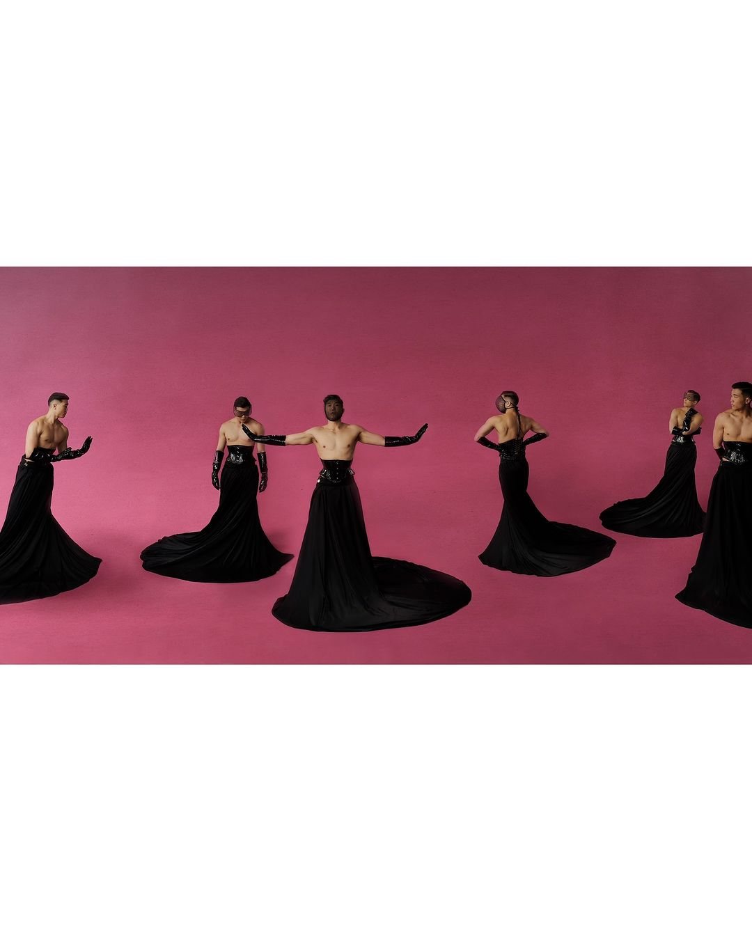 Group of seven people wearing black gowns and gloves pose against a solid pink background in a studio fashion photograph.