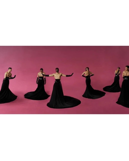 Group of seven people wearing black gowns and gloves pose against a solid pink background in a studio fashion photograph.