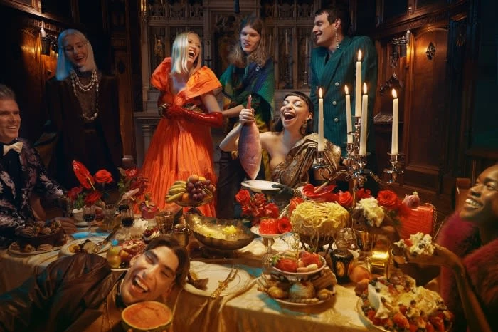 Festive indoor gathering with a diverse group of laughing people around a table laden with food, flowers, and candles in a dark, rich setting.