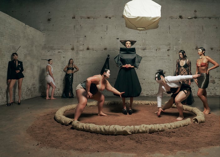 Group of women in an indoor setting, including two in sumo wrestling poses within a ring, surrounded by other women in fashionable clothing.