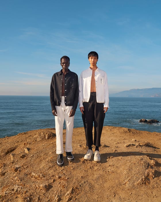 Two models pose on a coastal bluff overlooking the ocean under a blue sky. They wear fashionable jackets and trousers.