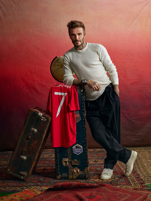 David Beckham poses with a soccer jersey and vintage luggage against a red backdrop. He wears casual attire and white sneakers.