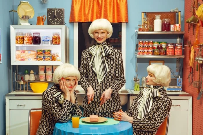 Three fair-skinned adult women in patterned dresses in a retro-style kitchen cutting a slice of pink cake on a blue tablecloth.