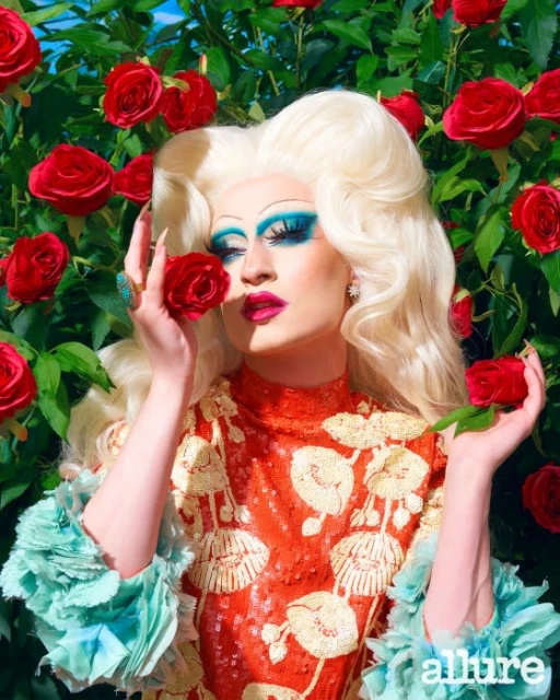 Drag queen poses in front of roses wearing a sequined gown with dramatic eye makeup and an ornate blonde wig.