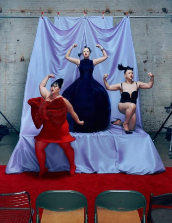 Three plus-size women strike poses in a studio setting. One wears a red dress with an oversized bow, another a blue gown, and another a black bodysuit.