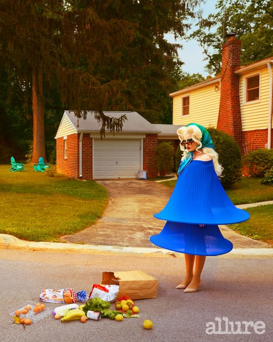 A person in an avant-garde blue outfit stands in a suburban street, surrounded by scattered groceries, with a house in the background.