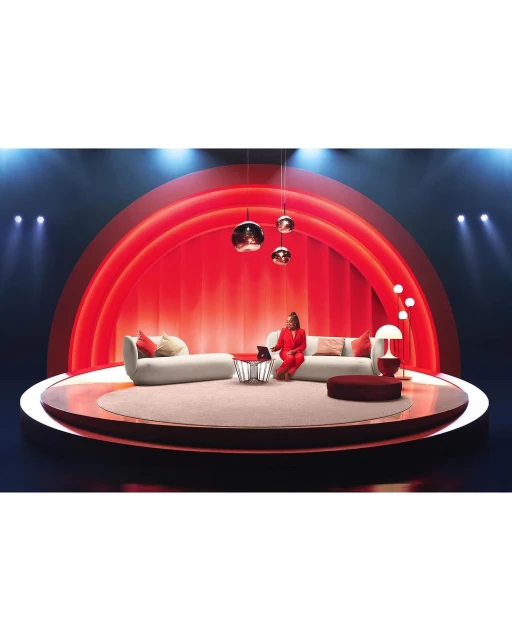Woman in a red suit sits on a couch on a circular stage, working on a laptop under hanging lights, spotlight and red background.