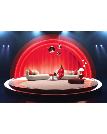 Woman in a red suit sits on a couch on a circular stage, working on a laptop under hanging lights, spotlight and red background.
