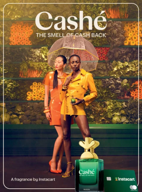 Two fashion models pose with a perfume bottle and Instacart card against a backdrop of colorful produce shelves.