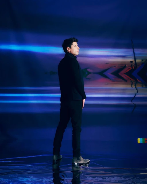 Adult male in dark clothing stands sideways against blue abstract background in studio lighting, creating a modern and sleek visual.