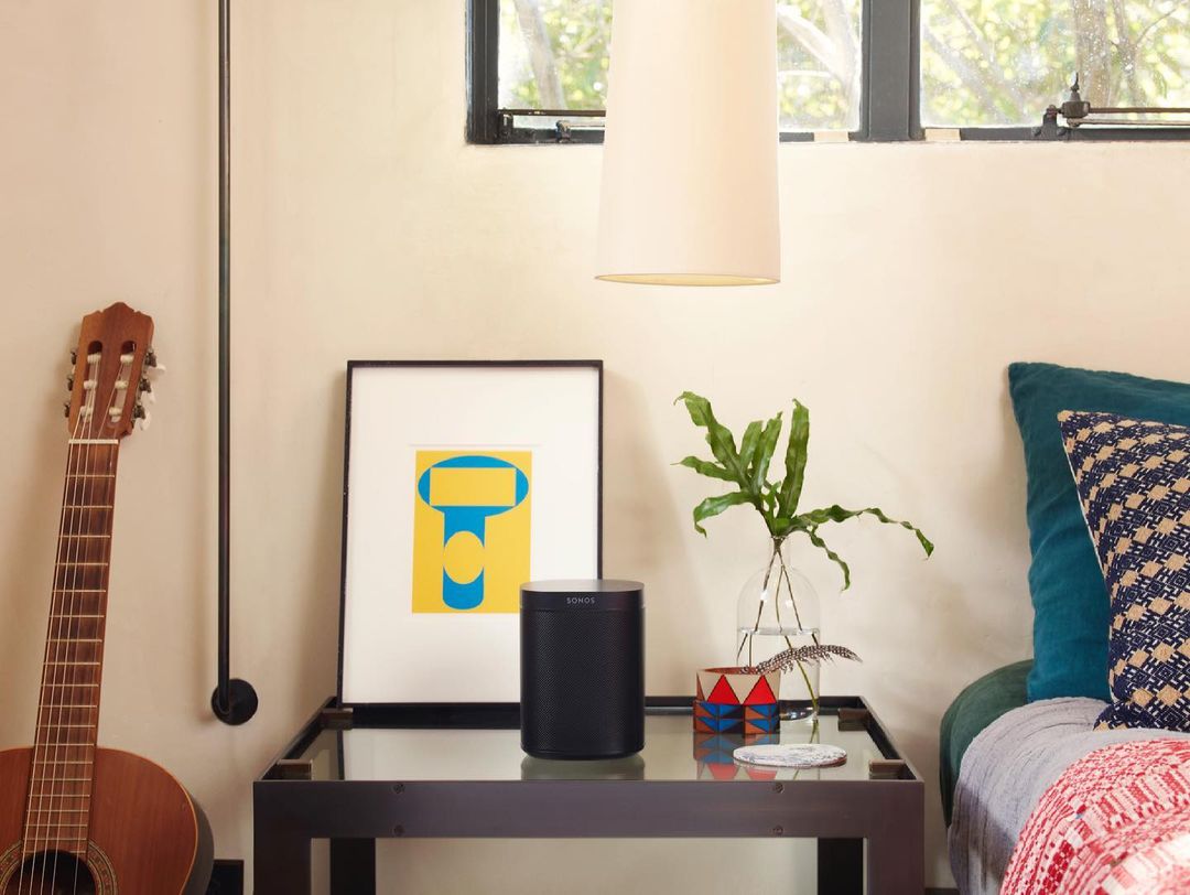 Living room setting featuring a SONOS speaker on a glass table, accented by art, a guitar, and lush greenery, creating a cozy, modern ambiance.