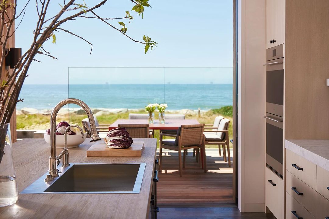 Modern coastal home interior showcases a kitchen with stainless steel sink, white cabinets, and ocean view through sliding glass doors.