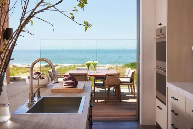 Modern coastal home interior showcases a kitchen with stainless steel sink, white cabinets, and ocean view through sliding glass doors.