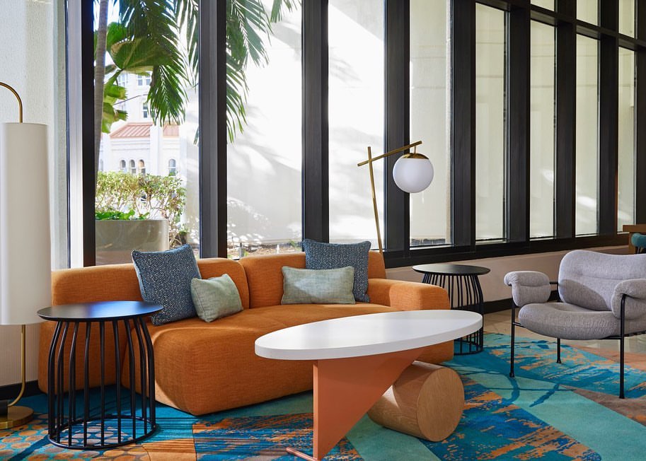 Lounge area with orange couch, blue pillows, round coffee table, and gray chair, with natural light streaming through large windows.
