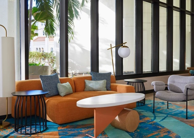 Lounge area with orange couch, blue pillows, round coffee table, and gray chair, with natural light streaming through large windows.