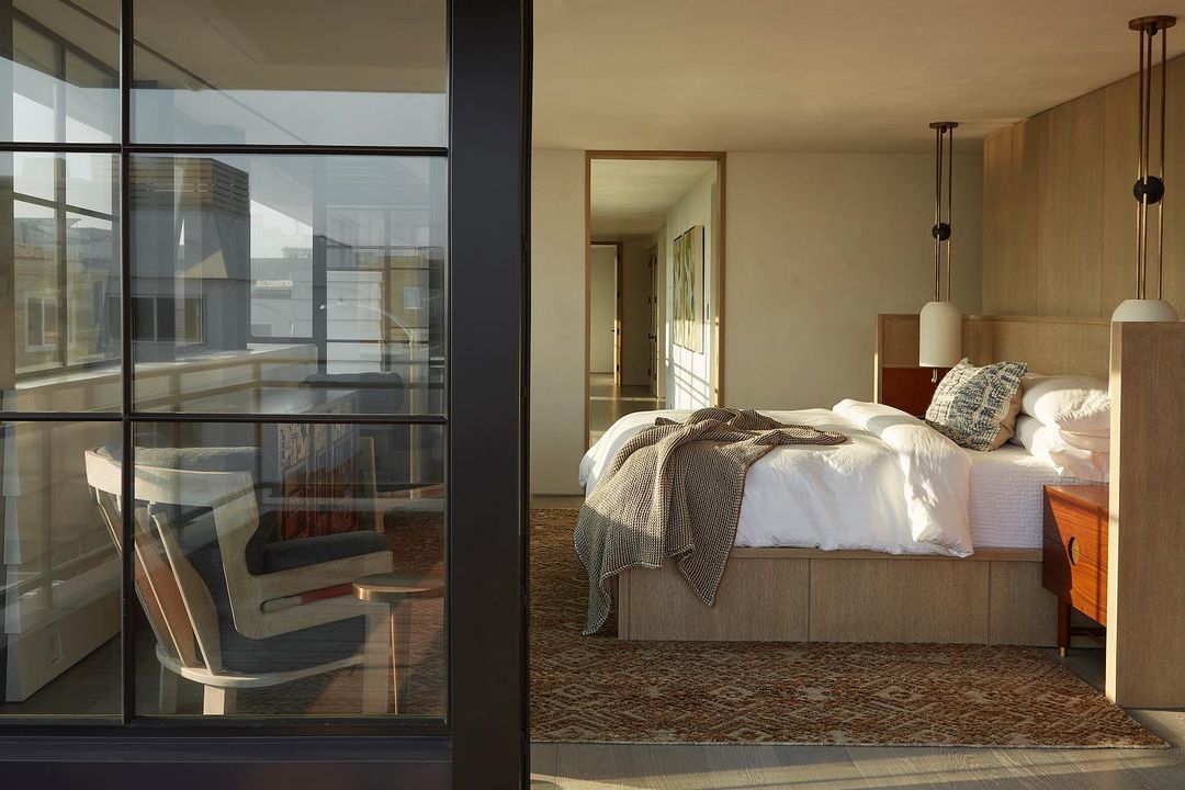 Bedroom interior featuring a bed with white linens, patterned throw, and bedside tables, viewed through a window framing an outdoor seating area.