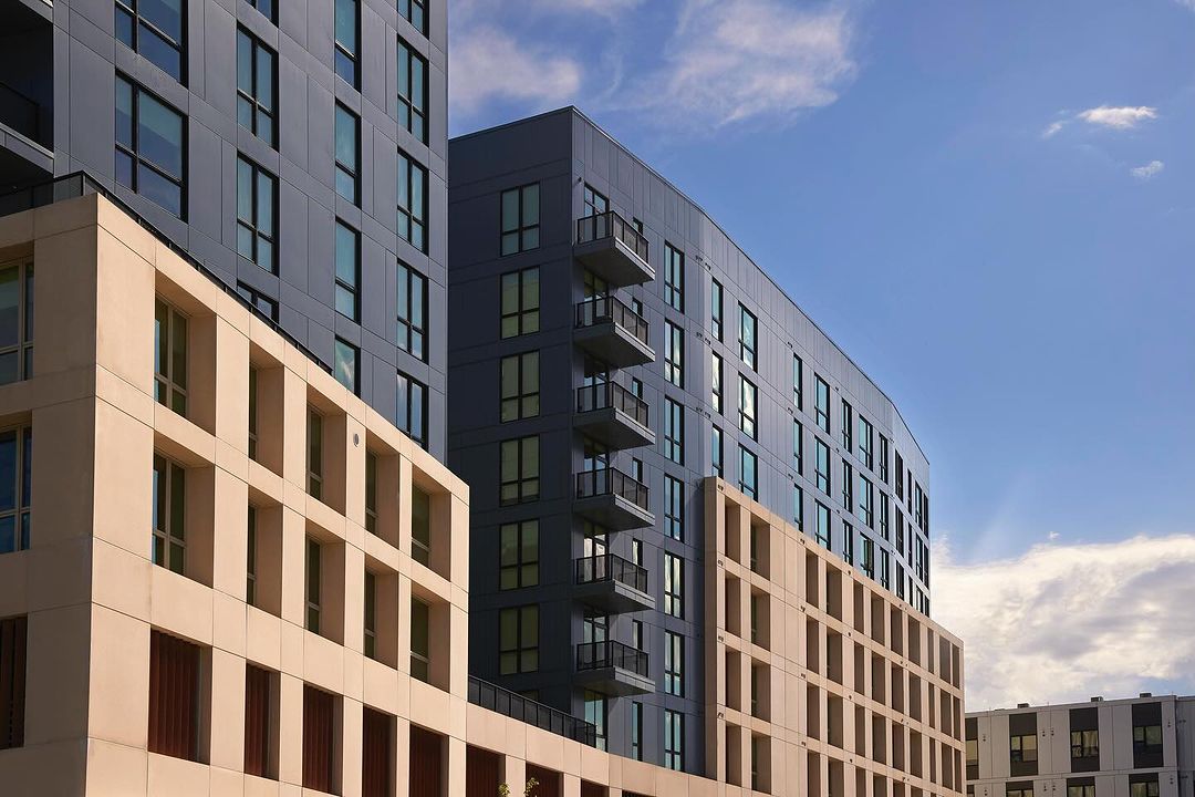 Exterior view of modern apartment buildings with gray and tan facades under a blue sky with scattered clouds, showcasing urban architecture.