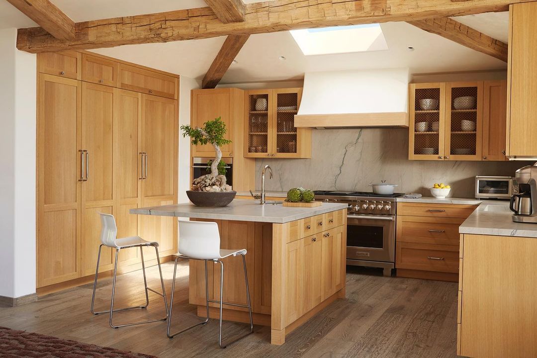 Bright kitchen interior with wood cabinets and island, featuring a bonsai tree and minimalist bar stools under wood beam ceiling.
