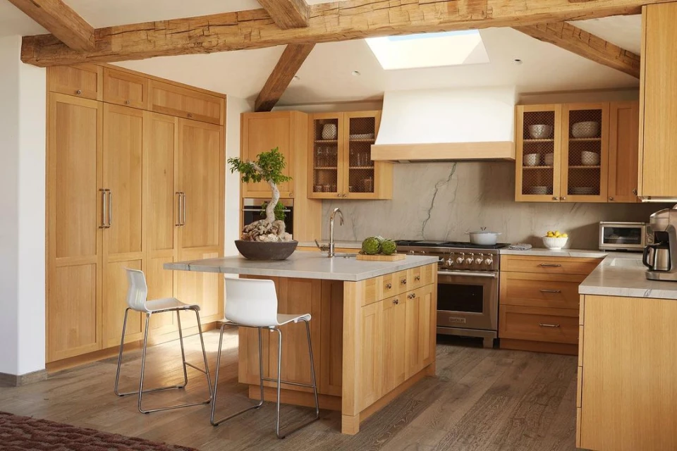Bright kitchen interior with wood cabinets and island, featuring a bonsai tree and minimalist bar stools under wood beam ceiling.