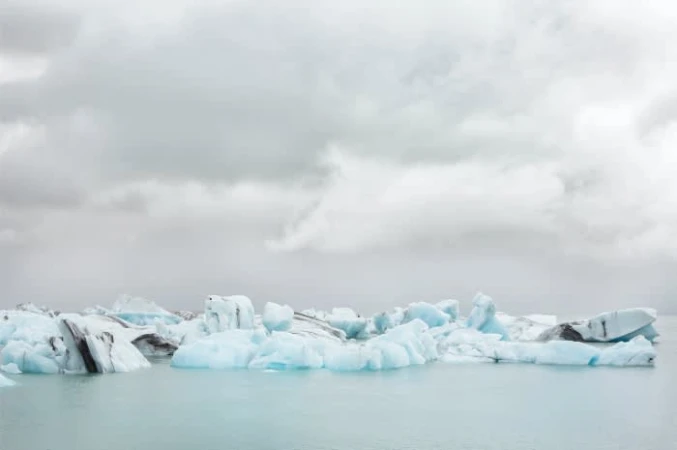Icebergs float on glacial water under an overcast sky, creating a serene and icy landscape scene with cool tones.