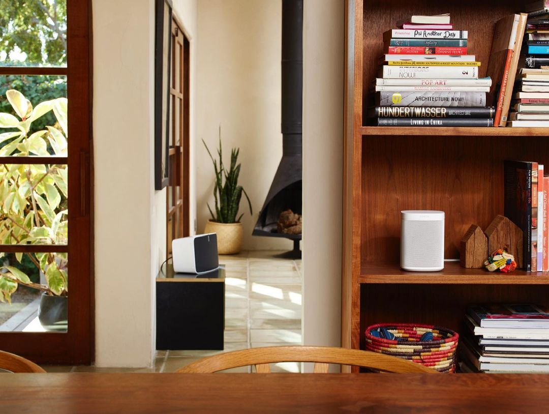 Indoor living room scene with two portable speakers in white, a wooden bookshelf, a fireplace, a dining table, and warm lighting.