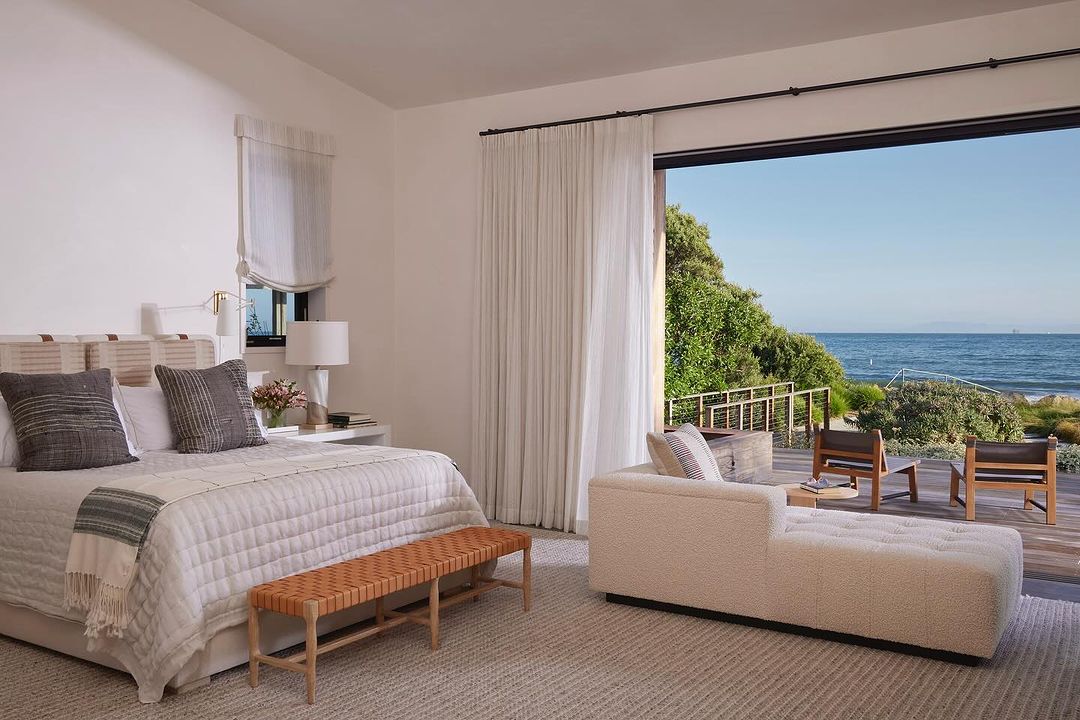 Bedroom featuring a bed, bench, and chaise lounge with an open doorway to a wooden deck overlooking the ocean and trees.