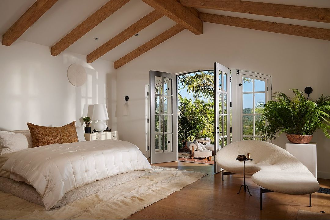 Bright, airy bedroom interior with white walls, wooden ceiling beams, and a view to a lush garden through open French doors.
