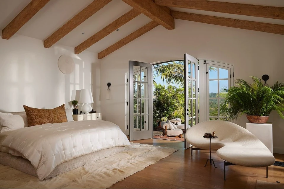 Bright, airy bedroom interior with white walls, wooden ceiling beams, and a view to a lush garden through open French doors.