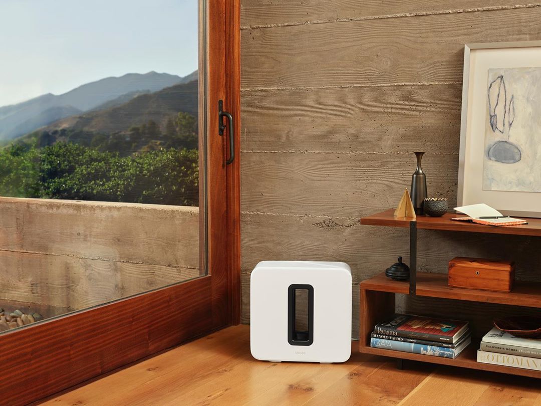 Interior shot of a white speaker on a wooden floor, with a window showing a mountain view and a wooden shelf with objects.